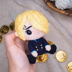 May include: A handmade crochet doll of a character with blonde hair, wearing a navy blue jacket with gold buttons. The doll has a stern expression and is surrounded by gold coins. The text "BOSQUEGURUMI" is visible in the upper left corner.
