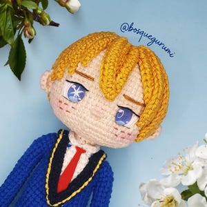 Może przedstawiać: Lalka szydełkowa z blond włosami, niebieskimi oczami i czerwonym krawatem. Lalka ma na sobie niebieską kurtkę ze złotą lamówką i białą koszulę. Lalka jest na jasnoniebieskim tle.