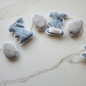 May include: A blue and white floral patterned fabric garland with bunny and egg shapes. The garland has a white border and is strung on a natural twine. The bunny shape has the name "Dominic" written on it.