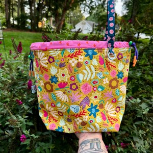 Puede incluir: Una bolsa de tela con cord&oacute;n ajustable sobre una base de color amarillo mostaza con un colorido estampado floral en tonos rosa, azul y blanco. La bolsa tiene un borde superior rosa brillante con estrellas, un asa azul con caritas sonrientes y cordones morados.