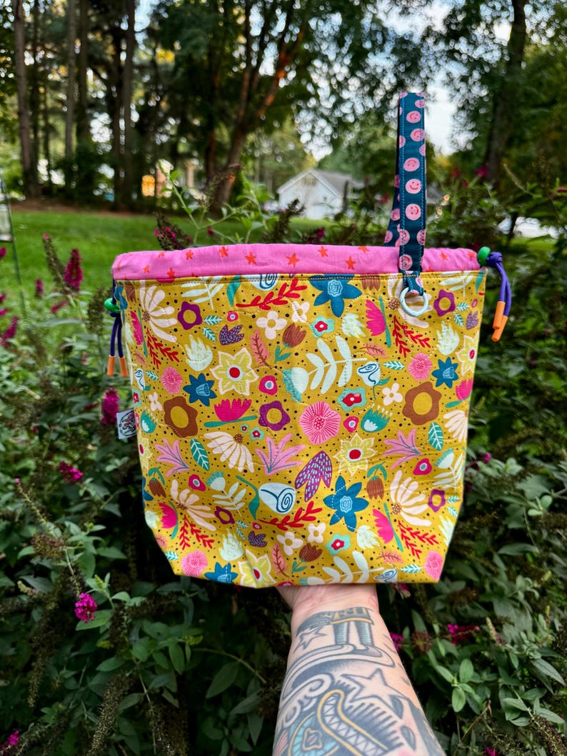 Puede incluir: Una bolsa de tela con cord&oacute;n ajustable sobre una base de color amarillo mostaza con un colorido estampado floral en tonos rosa, azul y blanco. La bolsa tiene un borde superior rosa brillante con estrellas, un asa azul con caritas sonrientes y cordones morados.
