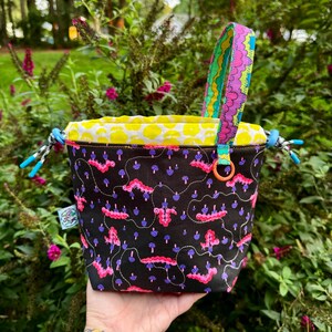 Puede incluir: Una bolsa con cord&oacute;n negra que presenta un patr&oacute;n de orugas rosas y peque&ntilde;os hongos azules. El bolso tiene un forro interior amarillo brillante con un estampado de hongos blancos, un asa colorida y cierres de cord&oacute;n azules en los laterales.