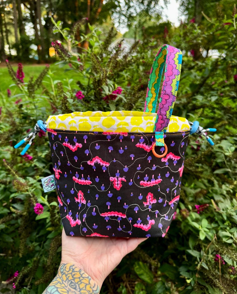 Puede incluir: Una bolsa con cord&oacute;n negra que presenta un patr&oacute;n de orugas rosas y peque&ntilde;os hongos azules. El bolso tiene un forro interior amarillo brillante con un estampado de hongos blancos, un asa colorida y cierres de cord&oacute;n azules en los laterales.