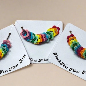 Könnte beinhalten: Drei handgefertigte girlanden in Blumenform in Regenbogenfarben, präsentiert auf weißen Karten. Jede Girlande besteht aus mehreren Filzblumen. Die Karten tragen die Aufschrift "FlockFest Fiber Arts".