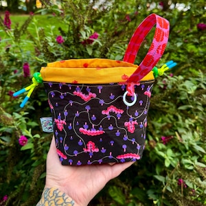 Puede incluir: Una bolsa con cord&oacute;n negra con un patr&oacute;n de orugas rosas y setas moradas, un forro interior amarillo brillante, un asa de tela con estampado rosa y cordones verde ne&oacute;n con puntas azules, rematada con una peque&ntilde;a anilla blanca.