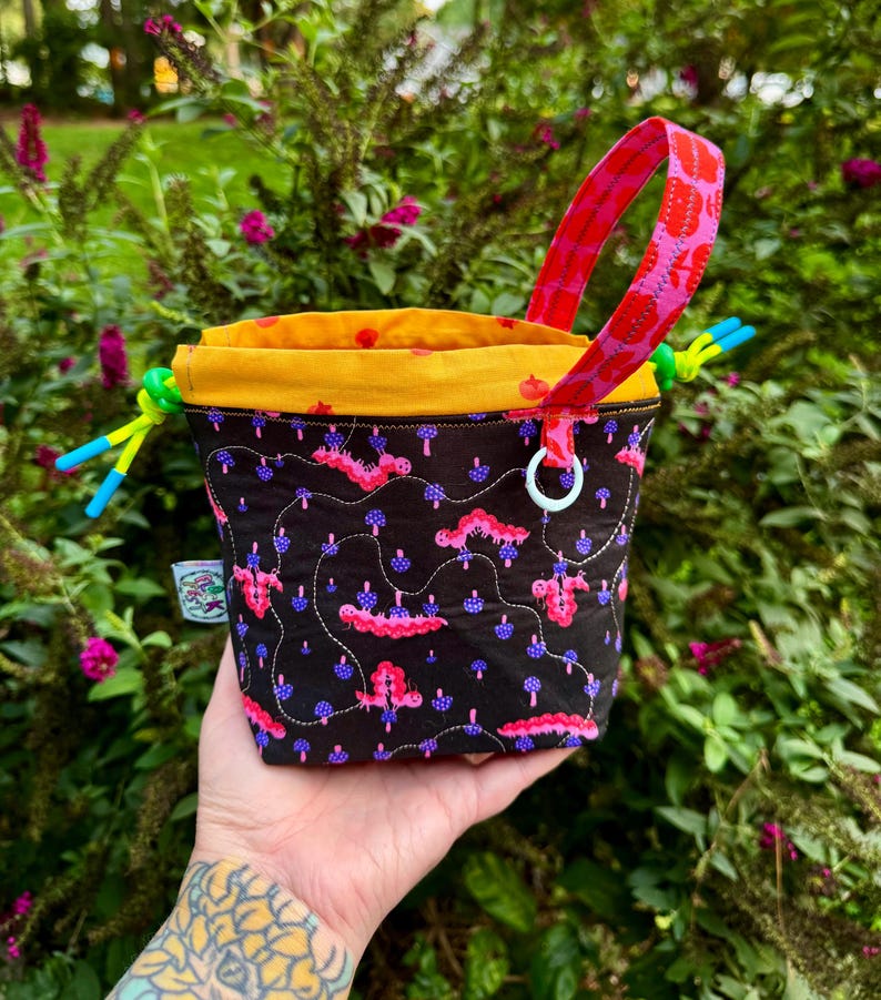 Puede incluir: Una bolsa con cord&oacute;n negra con un patr&oacute;n de orugas rosas y setas moradas, un forro interior amarillo brillante, un asa de tela con estampado rosa y cordones verde ne&oacute;n con puntas azules, rematada con una peque&ntilde;a anilla blanca.