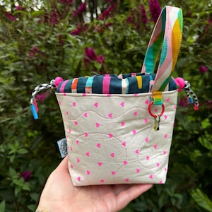 Puede incluir: Una peque&ntilde;a bolsa de tela con cord&oacute;n, exterior color crema con un patr&oacute;n de diminutos corazones rosa ne&oacute;n y costuras acolchadas onduladas. La parte superior es azul verdoso con rayas verticales coloridas, un asa arco&iacute;ris y cordones negros y blancos con topes de pl&aacute;stico azul y rosa.