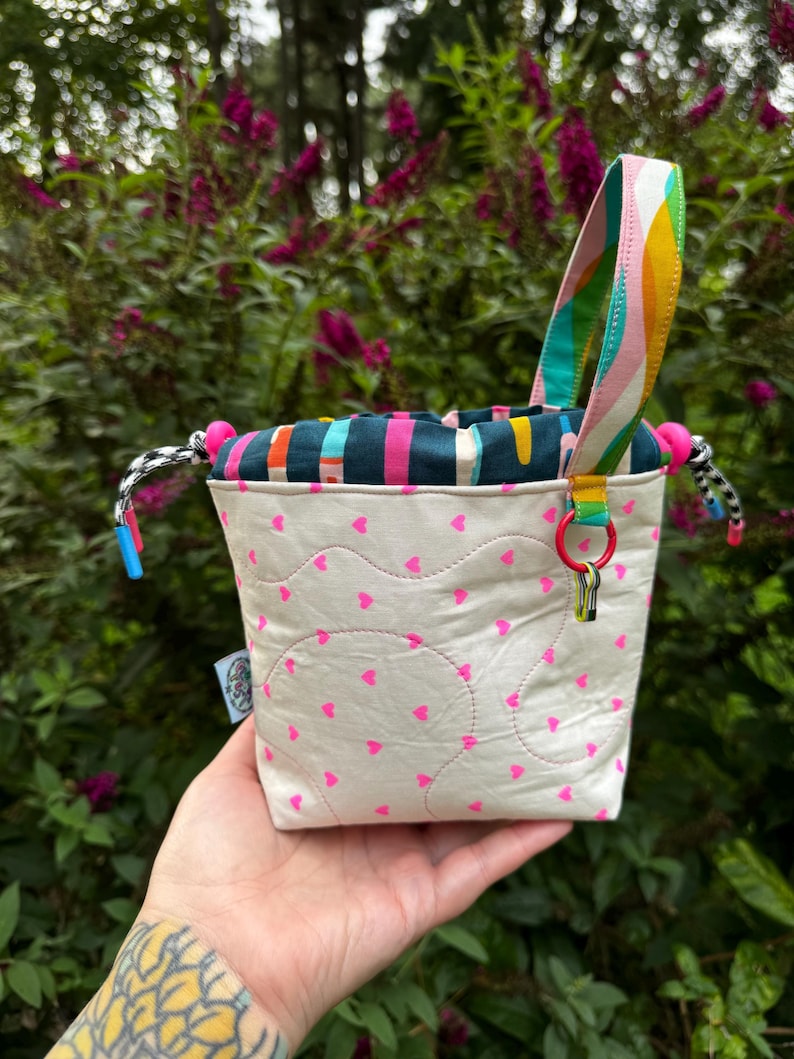 Puede incluir: Una peque&ntilde;a bolsa de tela con cord&oacute;n, exterior color crema con un patr&oacute;n de diminutos corazones rosa ne&oacute;n y costuras acolchadas onduladas. La parte superior es azul verdoso con rayas verticales coloridas, un asa arco&iacute;ris y cordones negros y blancos con topes de pl&aacute;stico azul y rosa.