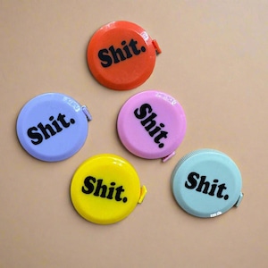 May include: A collection of five colorful, round tape measures. Each tape measure is a different color: orange, lavender, pink, yellow, and teal. The word "Shit." is printed in black on each tape measure.
