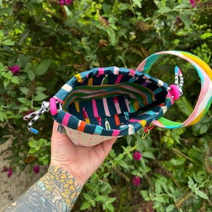Puede incluir: Una peque&ntilde;a bolsa de tela con cord&oacute;n que presenta un forro interior de color verde azulado oscuro con un patr&oacute;n de rayas verticales de colores. El exterior es de color crema con peque&ntilde;os tri&aacute;ngulos rosas, asa arco&iacute;ris y cierres rosas.