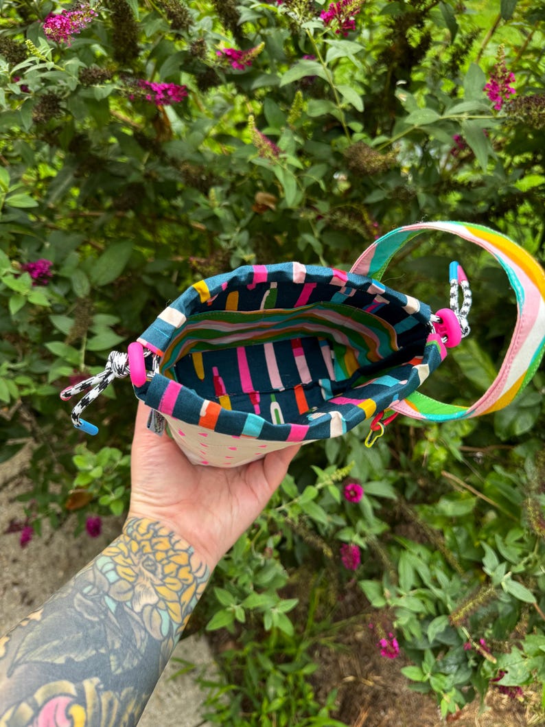 Puede incluir: Una peque&ntilde;a bolsa de tela con cord&oacute;n que presenta un forro interior de color verde azulado oscuro con un patr&oacute;n de rayas verticales de colores. El exterior es de color crema con peque&ntilde;os tri&aacute;ngulos rosas, asa arco&iacute;ris y cierres rosas.