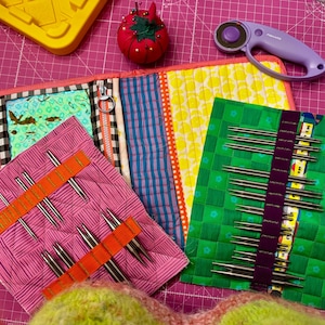 May include: A quilted fabric organizer case with a zipper, containing two smaller fabric needle cases with orange and purple elastic bands holding multiple metal knitting needles. A red tomato-shaped pincushion and a purple rotary cutter are nearby.
