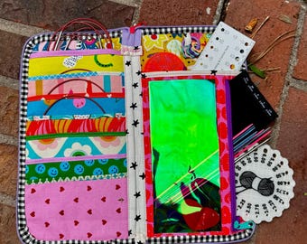 Puede incluir: Un estuche organizador de tela con múltiples bolsillos de patrones coloridos, incluyendo caras sonrientes, corazones y flores. El interior contiene agujas de tejer circulares, un medidor de agujas blanco y una herramienta redonda para hilos.