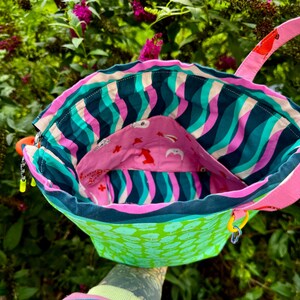 Puede incluir: Un bolso tipo cubo de tela con un exterior verde brillante con un patr&oacute;n de hojas repetitivo. El interior est&aacute; forrado con una tela de rayas onduladas en azul verdoso, azul oscuro y rosa, e incluye un bolsillo rosa decorado con peque&ntilde;os motivos animales rojos y blancos.