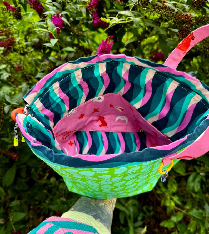 Puede incluir: Un bolso tipo cubo de tela con un exterior verde brillante con un patr&oacute;n de hojas repetitivo. El interior est&aacute; forrado con una tela de rayas onduladas en azul verdoso, azul oscuro y rosa, e incluye un bolsillo rosa decorado con peque&ntilde;os motivos animales rojos y blancos.
