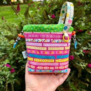 Puede incluir: Una bolsa de tela acolchada con cord&oacute;n que presenta un vibrante patr&oacute;n de rayas horizontales en tonos rosa, amarillo y turquesa. El bolso tiene un forro interior verde brillante, una correa estampada y cordones en blanco y negro con topes naranjas.