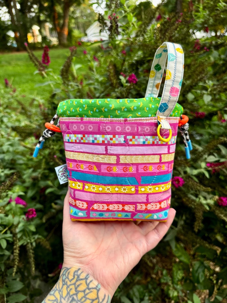 Puede incluir: Una bolsa de tela acolchada con cord&oacute;n que presenta un vibrante patr&oacute;n de rayas horizontales en tonos rosa, amarillo y turquesa. El bolso tiene un forro interior verde brillante, una correa estampada y cordones en blanco y negro con topes naranjas.