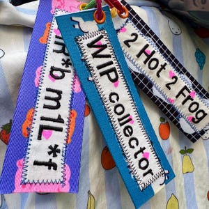 May include: Three rectangular fabric keychains attached to a metal ring. The purple one reads "r*b m1l*f", the blue one reads "WIP collector", and the black and white grid one reads "2 Hot 2 Frog". Each features embroidered text and pink hearts.