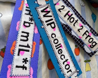 Puede incluir: Tres llaveros de tela rectangulares unidos a una anilla metálica. El morado dice "r*b m1l*f", el azul "WIP collector" y el de cuadrícula blanca y negra "2 Hot 2 Frog". Cada uno presenta texto bordado y corazones de color rosa.