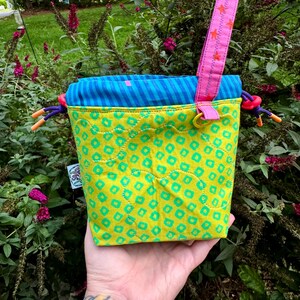Puede incluir: Una bolsa acolchada con cord&oacute;n de tela verde lima con un patr&oacute;n repetitivo de cuadrados verde azulado. La parte superior es de tela a rayas azules y verde azulado. Incluye una correa de mu&ntilde;eca rosa con estrellas naranjas y cordones morados.