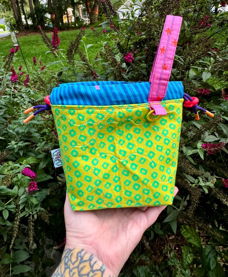 Puede incluir: Una bolsa acolchada con cord&oacute;n de tela verde lima con un patr&oacute;n repetitivo de cuadrados verde azulado. La parte superior es de tela a rayas azules y verde azulado. Incluye una correa de mu&ntilde;eca rosa con estrellas naranjas y cordones morados.