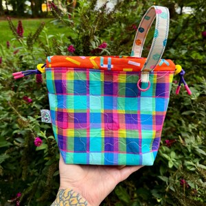 Puede incluir: Una bolsa acolchada con cord&oacute;n que presenta un vibrante patr&oacute;n de cuadros multicolor en tonos azul, morado, verde y amarillo. El bolso tiene un forro interior naranja con estampado geom&eacute;trico, un asa de tela estampada y cordones morados.