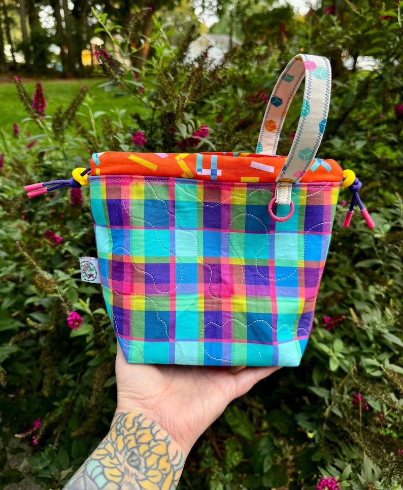 Puede incluir: Una bolsa acolchada con cord&oacute;n que presenta un vibrante patr&oacute;n de cuadros multicolor en tonos azul, morado, verde y amarillo. El bolso tiene un forro interior naranja con estampado geom&eacute;trico, un asa de tela estampada y cordones morados.