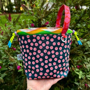 Puede incluir: Un bolso tipo cubo de tela con un exterior azul verdoso oscuro que presenta un patr&oacute;n repetitivo de peque&ntilde;as caras sonrientes rosas y l&iacute;neas blancas cosidas. El bolso tiene un borde superior ondulado colorido, un asa rosa brillante y cordones amarillo ne&oacute;n.