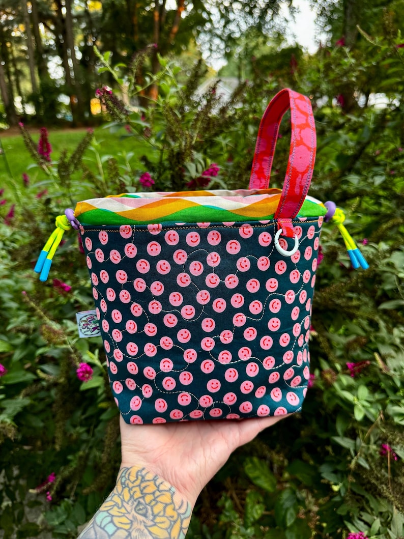Puede incluir: Un bolso tipo cubo de tela con un exterior azul verdoso oscuro que presenta un patr&oacute;n repetitivo de peque&ntilde;as caras sonrientes rosas y l&iacute;neas blancas cosidas. El bolso tiene un borde superior ondulado colorido, un asa rosa brillante y cordones amarillo ne&oacute;n.