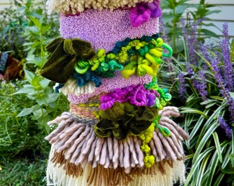 May include: A colorful wall hanging with a variety of textures and colors, including pink, green, purple, and brown. The hanging is made of yarn and fabric and has a fringe border.