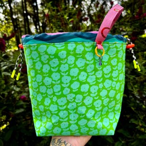 Puede incluir: Una bolsa con cord&oacute;n de tela verde brillante con un patr&oacute;n floral verde claro repetido. El borde superior tiene un dise&ntilde;o ondulado azul y azul verdoso, un asa rosa con motivos de arco&iacute;ris rojos y cordones blancos y negros con puntas amarillo ne&oacute;n.