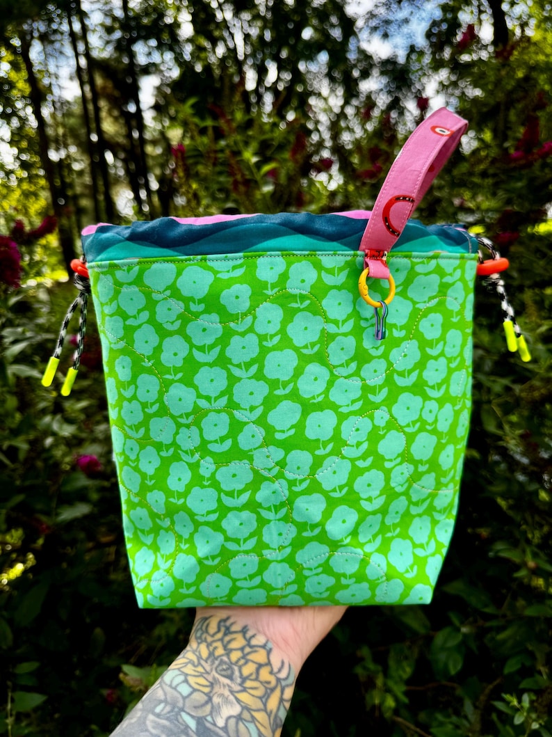 Puede incluir: Una bolsa con cord&oacute;n de tela verde brillante con un patr&oacute;n floral verde claro repetido. El borde superior tiene un dise&ntilde;o ondulado azul y azul verdoso, un asa rosa con motivos de arco&iacute;ris rojos y cordones blancos y negros con puntas amarillo ne&oacute;n.