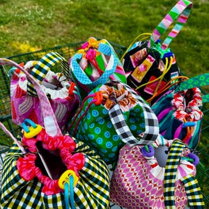 Puede incluir: Una colección de pequeñas bolsas de tela con cordón ajustable que presentan varios patrones coloridos, incluyendo caras sonrientes verdes, cuadros vichy en blanco y negro, formas geométricas rosas y estampados florales, cada una con asa.
