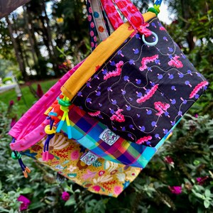 Puede incluir: Un conjunto de tres bolsas de tela con cord&oacute;n ajustables con patrones variados, incluyendo una bolsa negra con orugas rosas y setas azules, una bolsa a cuadros azul, verde y rosa, y una bolsa amarilla con motivos florales y el texto Fluff Fest.