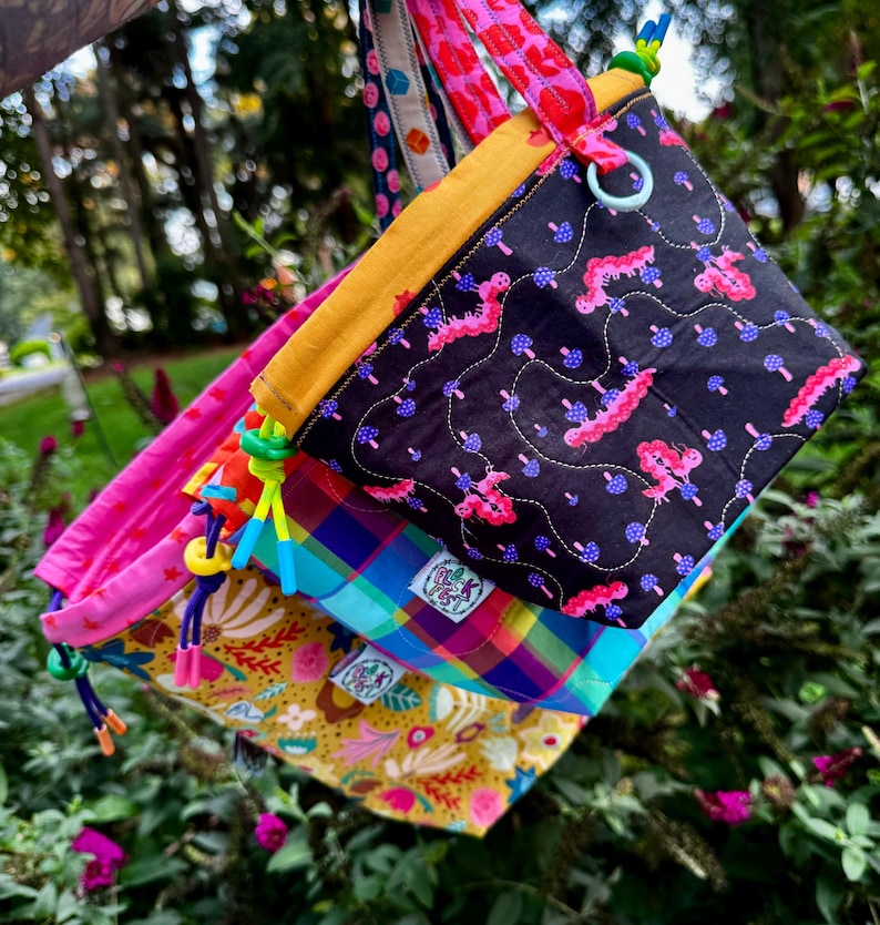 Puede incluir: Un conjunto de tres bolsas de tela con cord&oacute;n ajustables con patrones variados, incluyendo una bolsa negra con orugas rosas y setas azules, una bolsa a cuadros azul, verde y rosa, y una bolsa amarilla con motivos florales y el texto Fluff Fest.