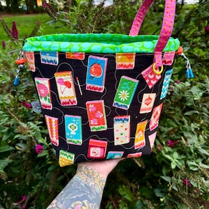 Puede incluir: Un bolso tipo cubo de tela con exterior negro que presenta un estampado colorido de cajas de cerillas con diversas ilustraciones. El bolso tiene un forro interior verde brillante, un asa estampada rosa y cierres de cord&oacute;n con topes azules y naranjas.