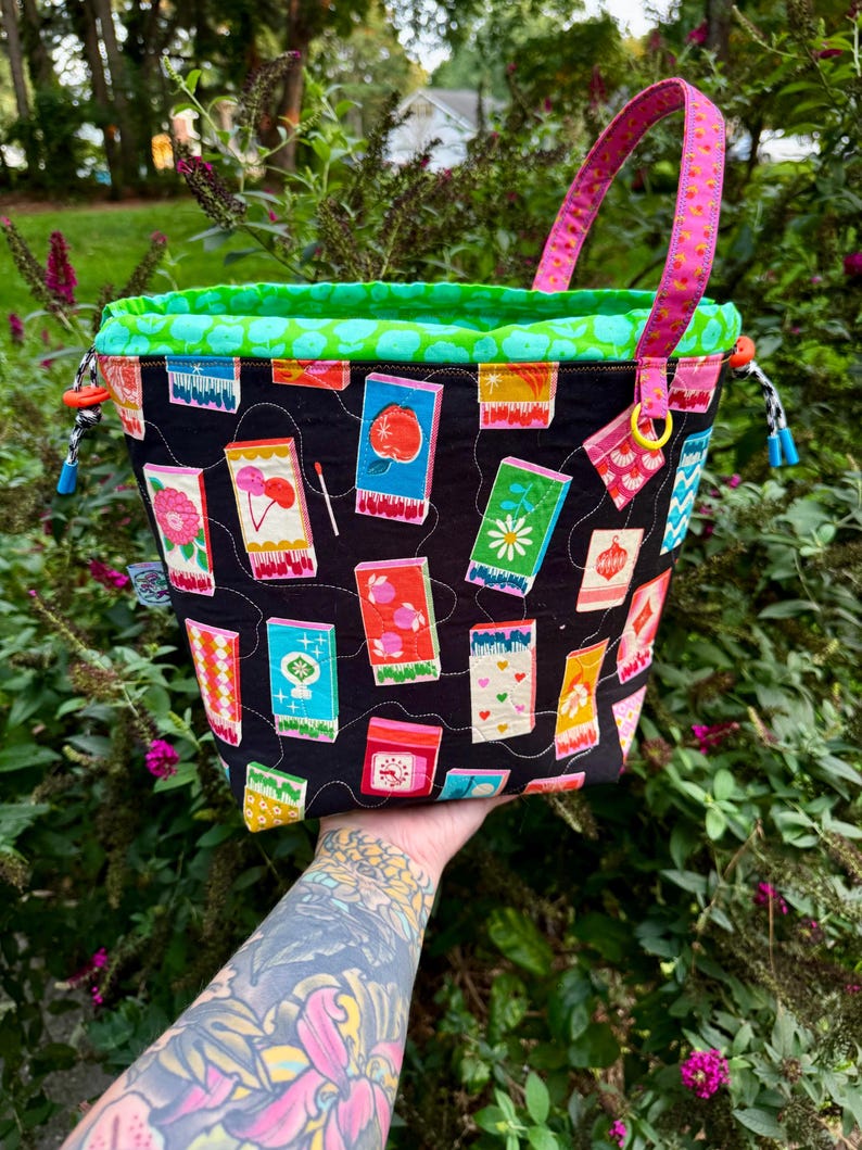 Puede incluir: Un bolso tipo cubo de tela con exterior negro que presenta un estampado colorido de cajas de cerillas con diversas ilustraciones. El bolso tiene un forro interior verde brillante, un asa estampada rosa y cierres de cord&oacute;n con topes azules y naranjas.