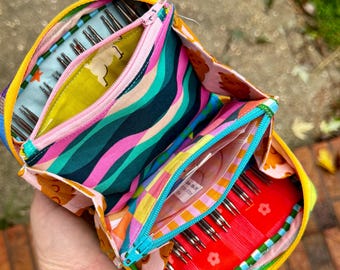Puede incluir: Un estuche organizador de tela con múltiples compartimentos y un patrón multicolor vibrante de líneas onduladas, flores y estampados de animales. Incluye dos secciones con cremallera que contienen varias agujas de tejer de metal y bolsas.