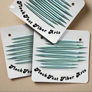 Puede incluir: Tres paquetes de agujas de tejer Flock Fest Fiber Arts. Cada paquete contiene varias agujas de tejer de color verde azulado claro. Los paquetes son blancos con el nombre de la marca impreso en negro. El fondo es de color beige claro.