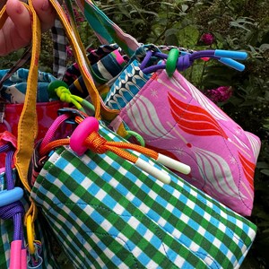 Puede incluir: Una colecci&oacute;n de bolsas de tela acolchada con cord&oacute;n ajustable que presentan varios patrones, incluyendo cuadros vichy verdes y azules, rosa con formas abstractas rojas y blancas, y azul con motivos animales blancos. Cada bolsa tiene un cord&oacute;n y un cierre de pl&aacute;stico.