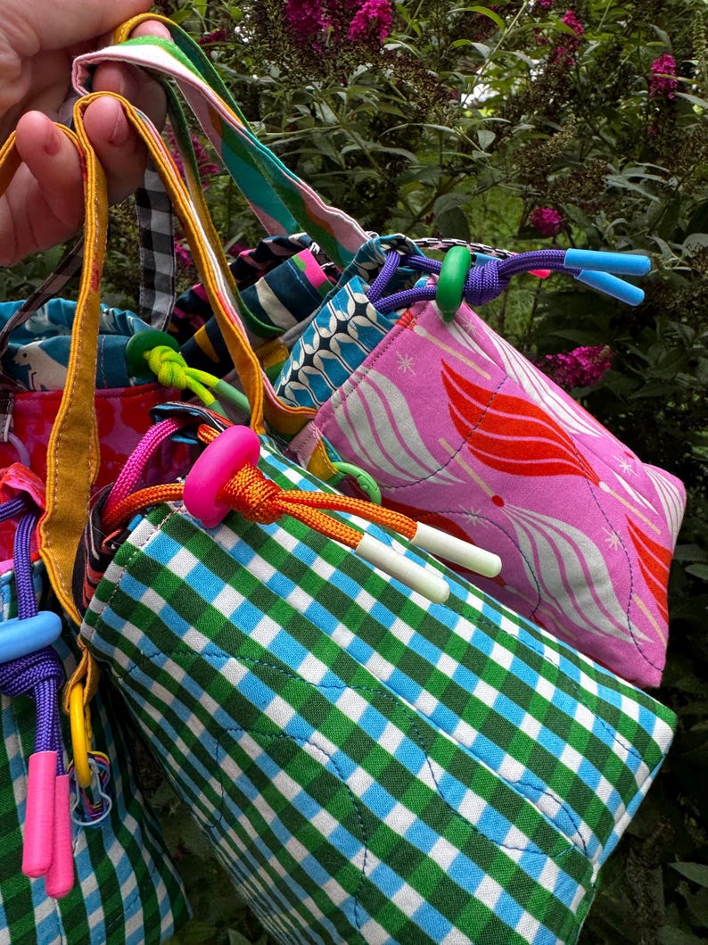 Puede incluir: Una colecci&oacute;n de bolsas de tela acolchada con cord&oacute;n ajustable que presentan varios patrones, incluyendo cuadros vichy verdes y azules, rosa con formas abstractas rojas y blancas, y azul con motivos animales blancos. Cada bolsa tiene un cord&oacute;n y un cierre de pl&aacute;stico.