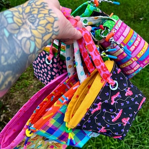 May include: A collection of small fabric drawstring bags featuring various colorful patterns including black with pink mushrooms, bright plaid, and geometric designs. Each bag has a fabric handle, a drawstring closure with plastic toggles.