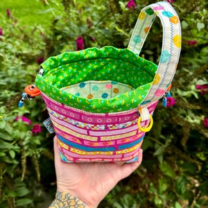 Puede incluir: Una bolsa de tela con cord&oacute;n que presenta un exterior rosa con bandas horizontales de patrones geom&eacute;tricos coloridos, un forro interior verde brillante con un peque&ntilde;o estampado floral y un asa blanca decorada con un motivo de cubos.