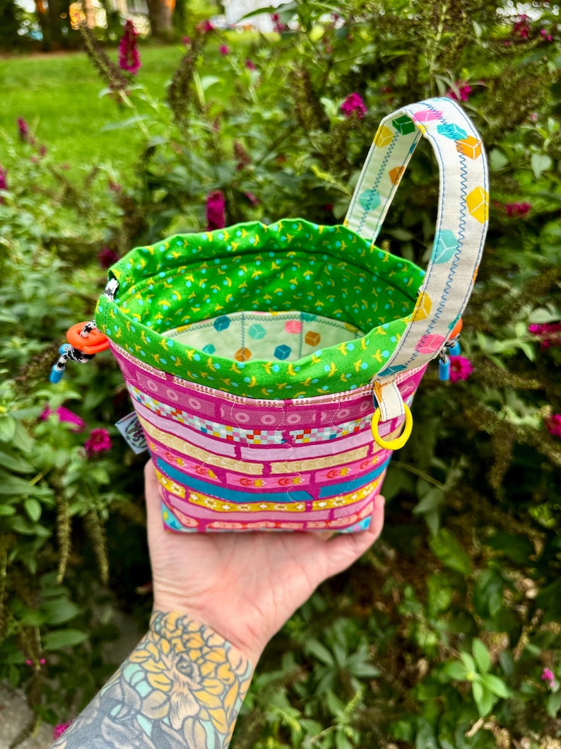 Puede incluir: Una bolsa de tela con cord&oacute;n que presenta un exterior rosa con bandas horizontales de patrones geom&eacute;tricos coloridos, un forro interior verde brillante con un peque&ntilde;o estampado floral y un asa blanca decorada con un motivo de cubos.
