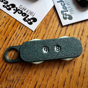 以下が含まれることがあります： 数字の9と8が表示された濃いティール色のステッチカウンター。カウンターには小さなループと2つの白いローラーが付いています。背景には「Flock Fest Fiber Arts」の文字が入ったカードがあります。