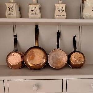 May include: Four copper saucepans with dark metal handles are arranged in a row. Above them, on a shelf, sit four cream-coloured ceramic canisters with floral designs and labels including the word SALT, displayed against a white wooden wall.