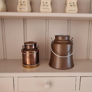 May include: Two metal milk churns sit on a shelf. The smaller churn is polished copper with a brass base and a wire handle. The larger churn is a matte brown metal with a silver-toned wire handle. Above them, a shelf holds five cream-coloured ceramic jars.