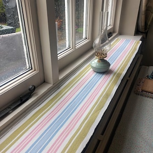 May include: A white windowsill with a striped runner in shades of green, pink, and blue. A glass oil lamp with a teal base sits on the runner.