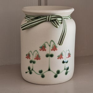May include: A cream-coloured ceramic jar with a green and white ribbon tied around the top. The jar is decorated with a floral design in red and green. The jar is a decorative item.