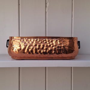 May include: An oblong, hammered copper planter with a metallic, reflective finish. The vessel features rounded corners and two small, black metal handles attached to the sides, providing a contrast to the warm, orange-toned copper surface.